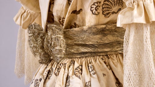 Close-up of the Cockleshell Dress, showing its cream patterned fabric, gathered waist and metallic sash with decorative bow.